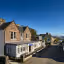 This is a well-composed exterior shot of a stone inn in a coastal village. The image is sharp with excellent depth of field, ensuring both the foreground building and the background scenery are in focus. The lighting is bright and sunny, providing natural, accurate colors. There are no unwanted elements present, and the architectural lines are straight and professional. The only minor drawback is the heavy shadow cast on the right side of the street, which creates a slight contrast imbalance, but overall it is a high-quality, realistic photograph.