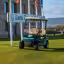 The image is well-composed with the golf cart as the clear focal point against the background of the clubhouse sign and flags. It is well-lit with natural sunlight, resulting in good color accuracy and depth. The image is sharp and displays high photo-realism. There are no unwanted elements such as logos, food, or clutter present. The focus is sharp on the golf cart, effectively capturing the essence of the location. Score: 85. Strengths: Excellent composition, clear subject focus, and natural, high-quality lighting. Weaknesses: Minor potential for a more dynamic angle, but overall very professional.